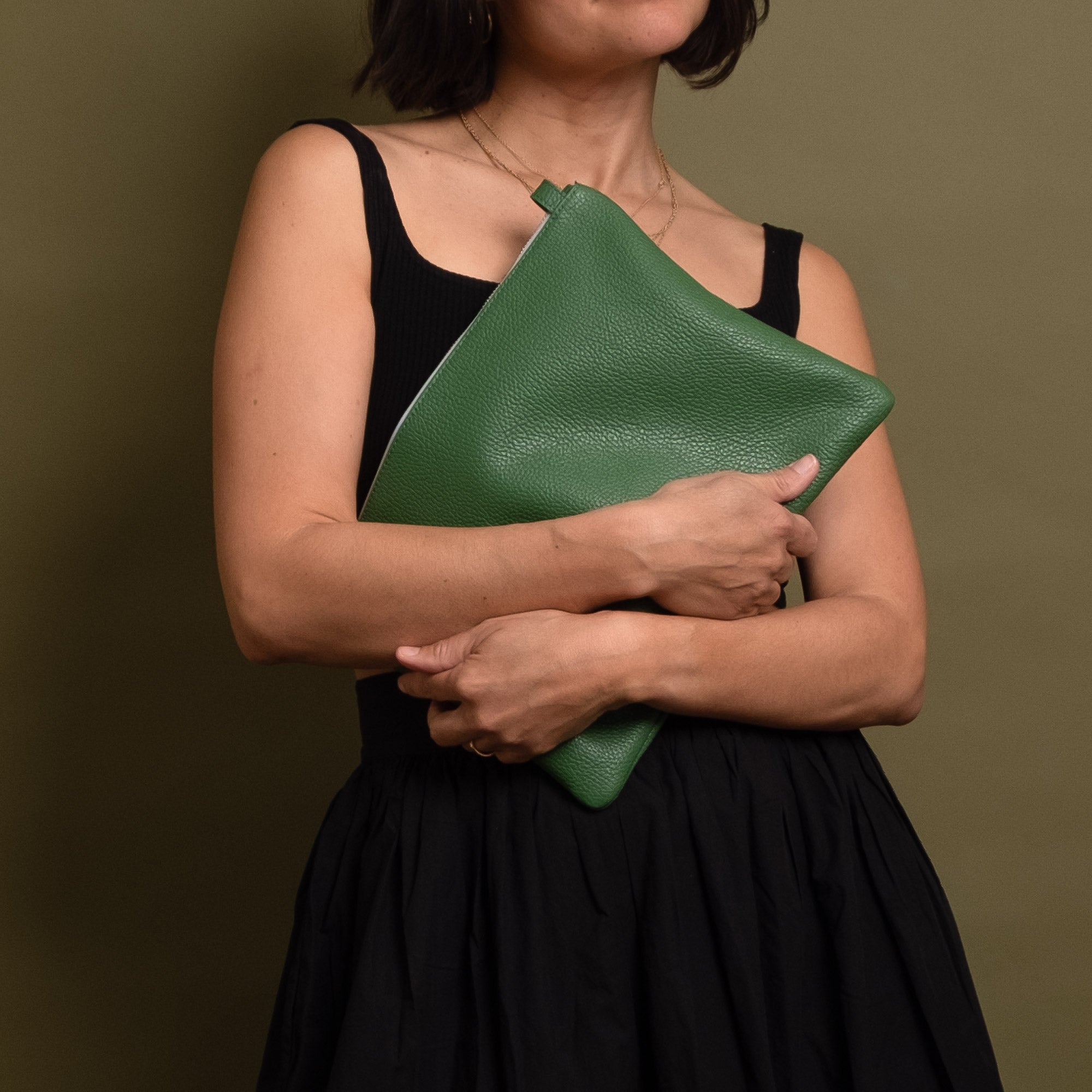 A woman in a black dress holds a green pebbled leather clutch bag against her torso, shown from the shoulders down against a solid olive-brown background.