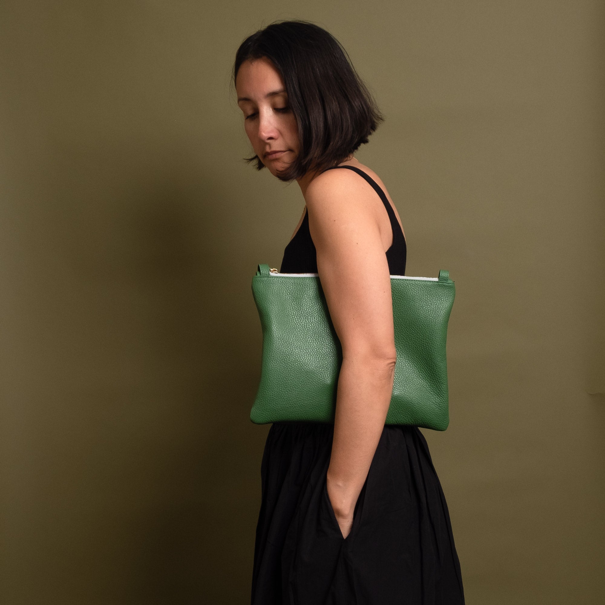 A woman in a black dress holds a sage green textured leather clutch bag against a solid olive green background.