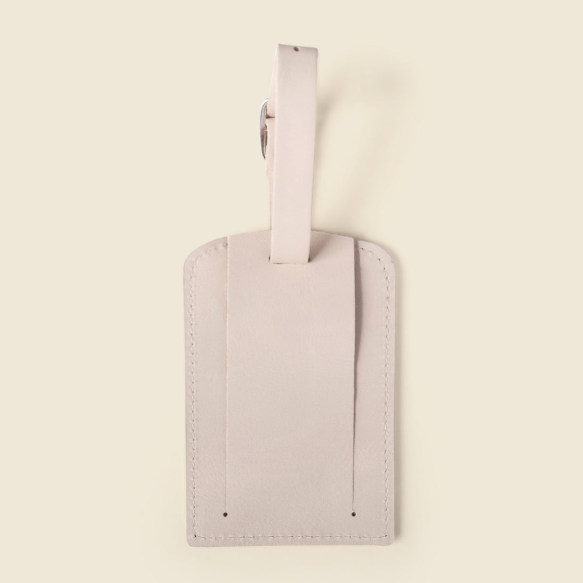 A minimalist light pink leather luggage tag with a matching strap and a small silver metal ring, shown against a solid cream background.