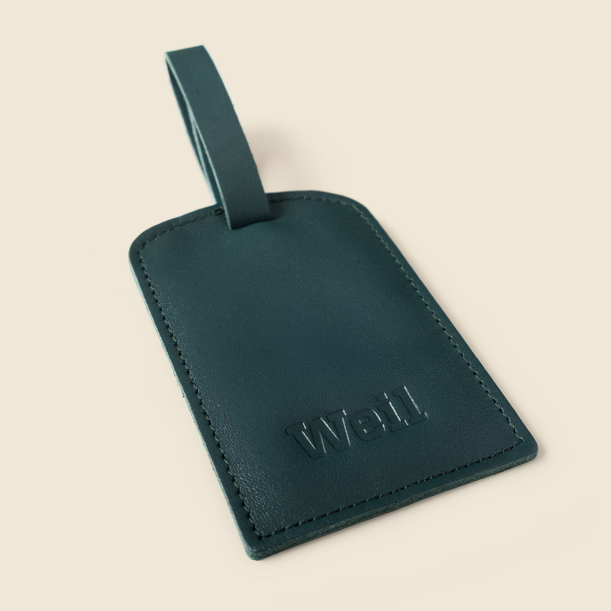 A dark teal leather luggage tag with a matching loop strap, featuring the debossed brand name 'We'll' on the front, set against a solid cream background.