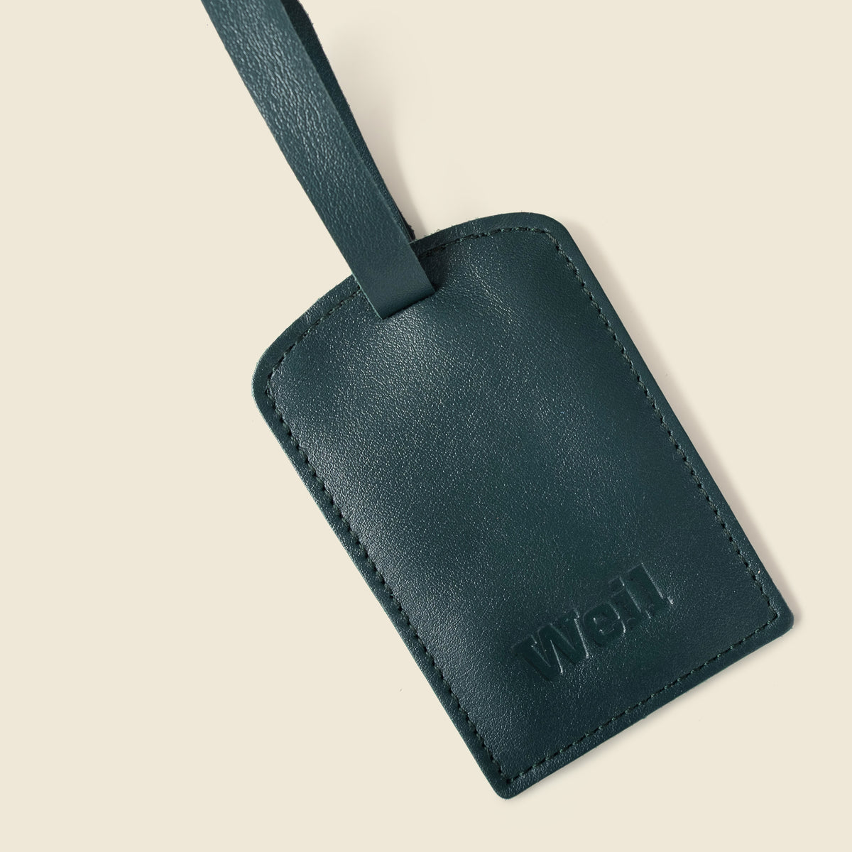 A dark teal leather luggage tag with a matching strap, featuring the debossed brand name 'wei.' in the bottom right corner, set against a solid cream background.
