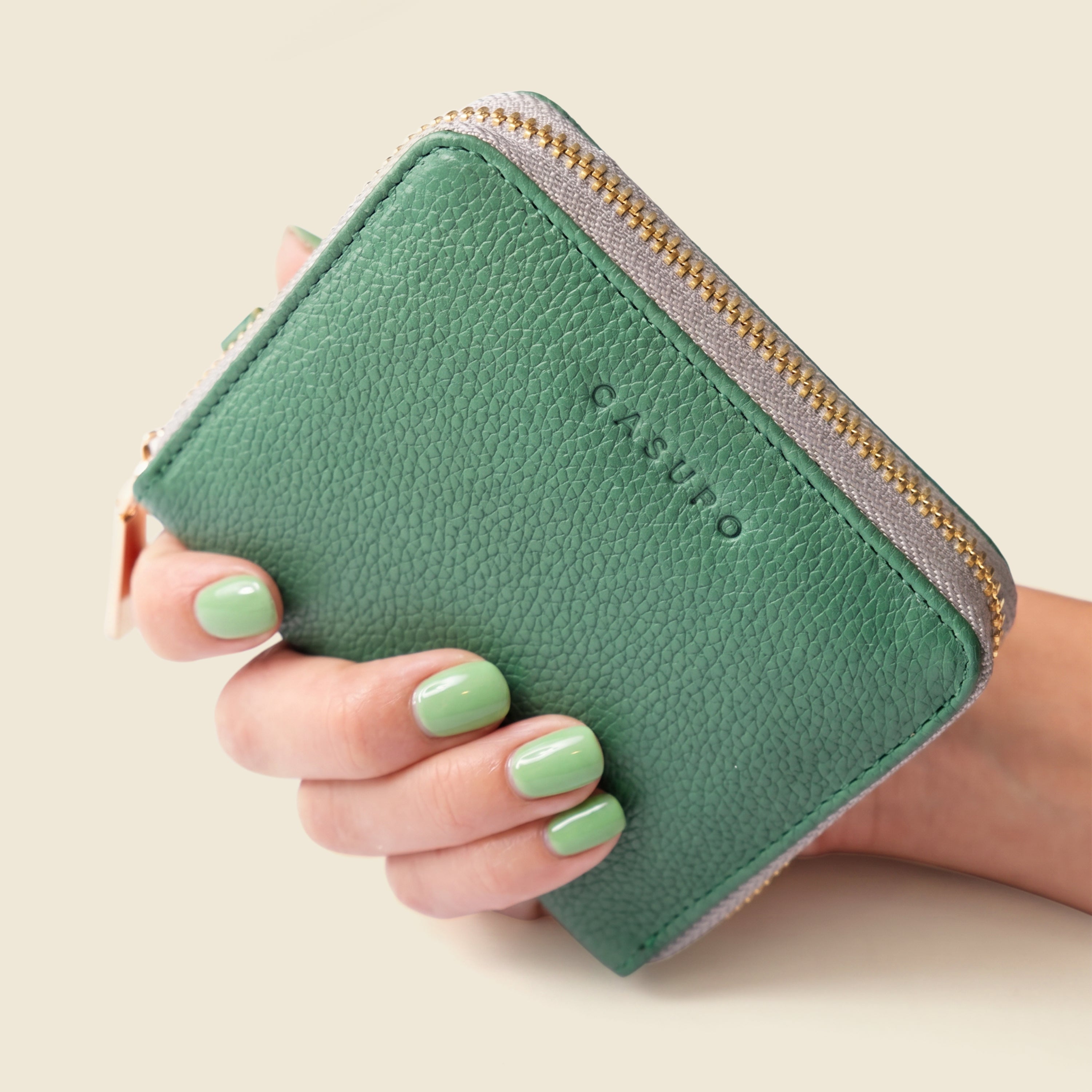 Green wallet with gold zipper held by a hand with green nail polish on a beige background