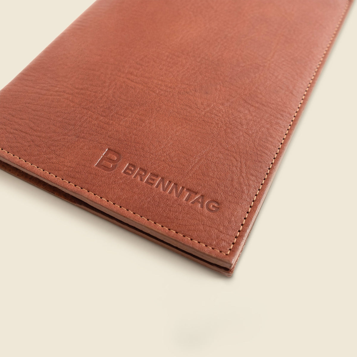 A close-up of a brown leather notebook cover or sleeve with visible stitching and an embossed Brenntag logo, photographed on a light cream background.