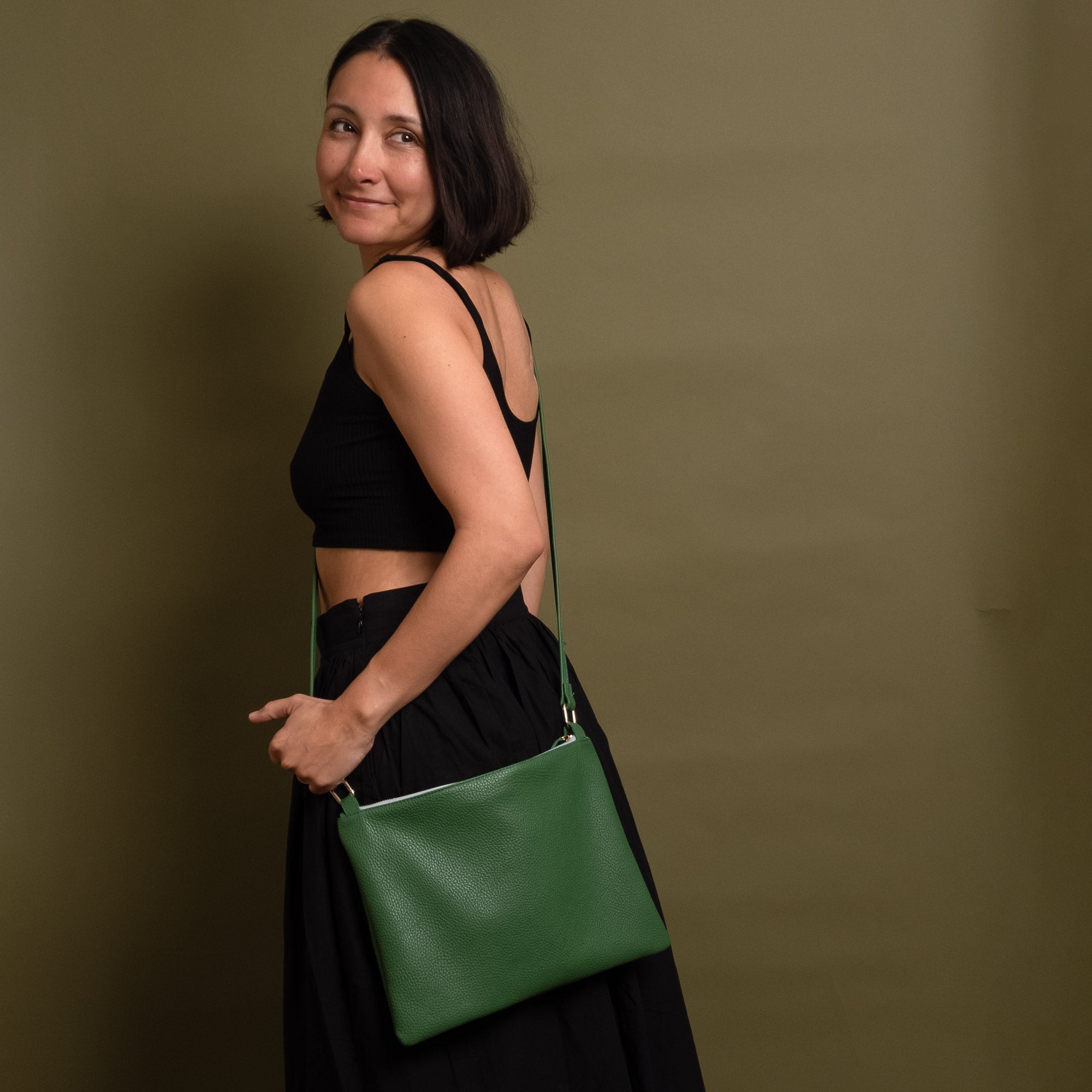 A woman wearing a black crop top and black skirt carries a green leather crossbody bag with a thin silver zipper, posing against a solid olive green background.