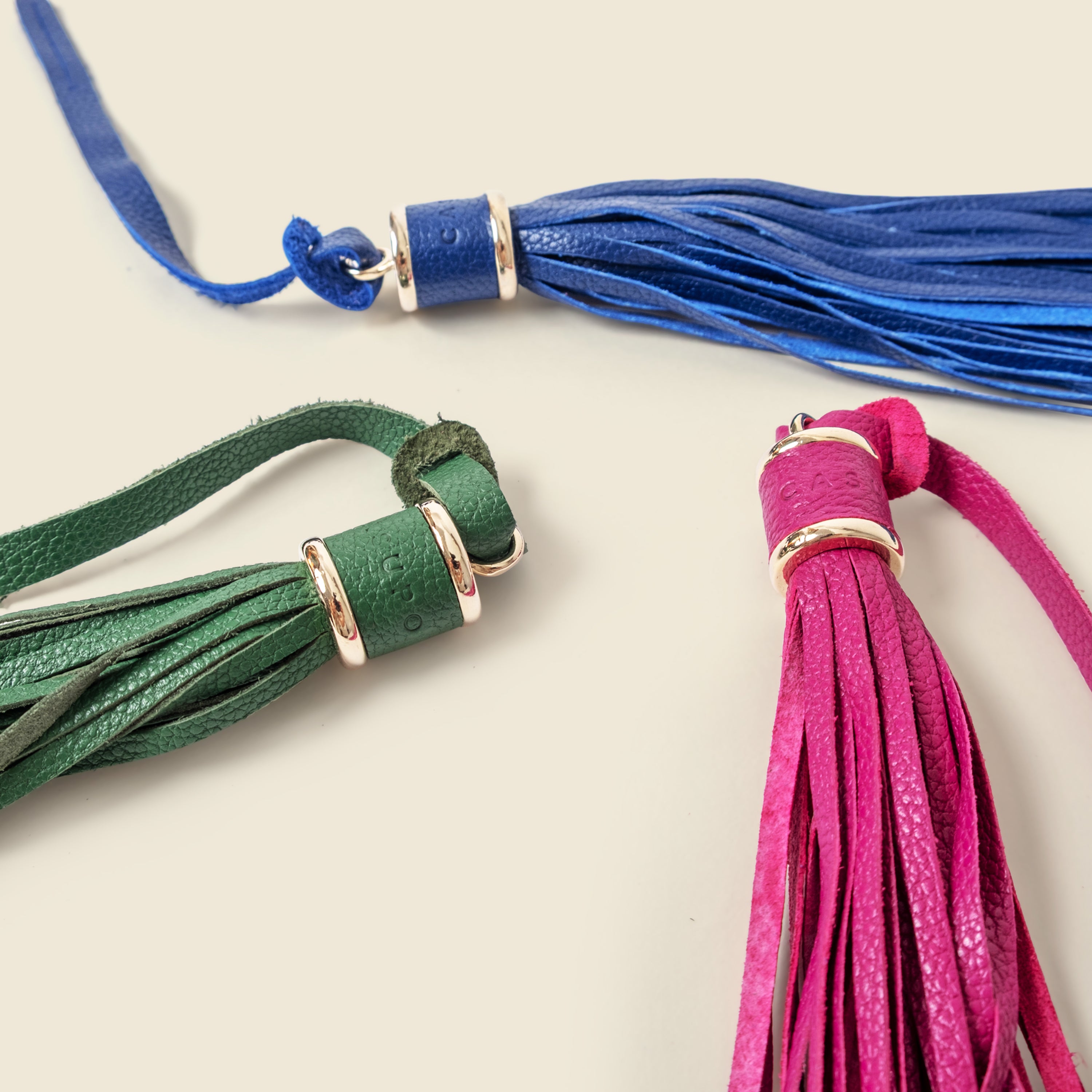 Three colorful tassels (blue, green, pink) with gold accents on a light background