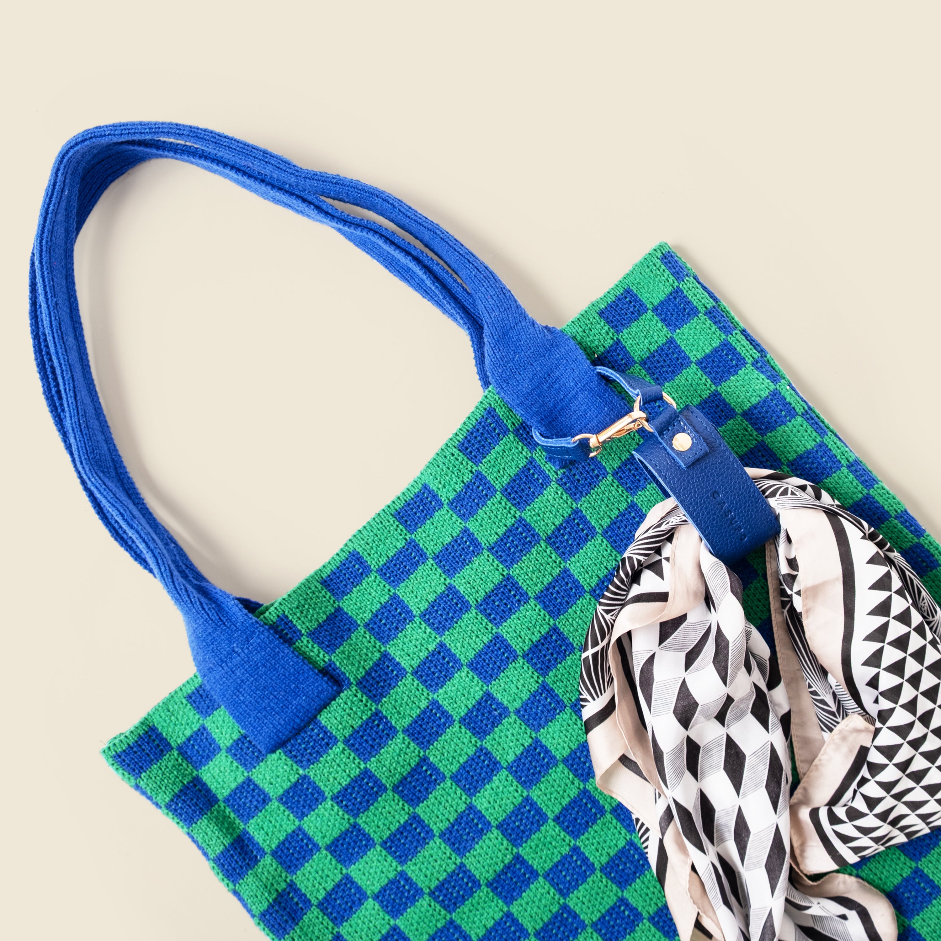 Green and blue checkered bag with a blue strap and a patterned scarf on a beige background
