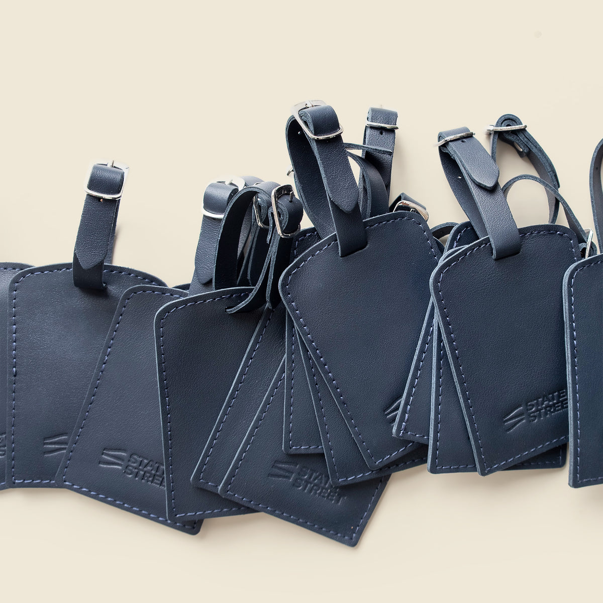 A row of dark navy blue leather luggage tags with matching straps and silver-tone buckles, arranged diagonally on a light beige background. 