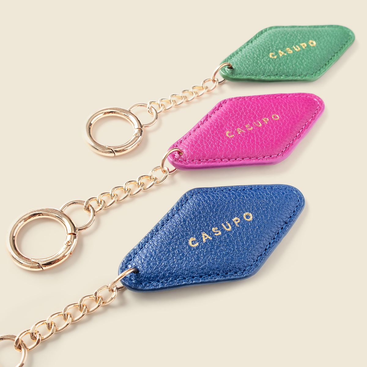 Three diamond-shaped leather keychains in green, pink, and blue, each with a gold-tone chain and key ring, displayed on a light beige background. The brand name 'CASUPO' is embossed on each piece.