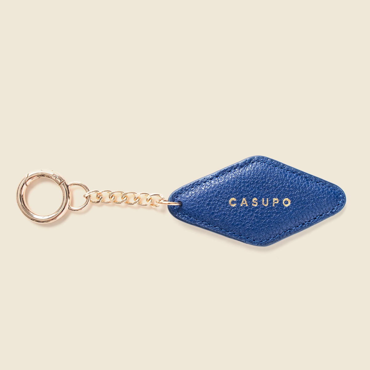 A blue textured leather keychain tag with a gold-tone chain and key ring, featuring the brand name 'CASUPO' in gold lettering, set against a solid cream background.