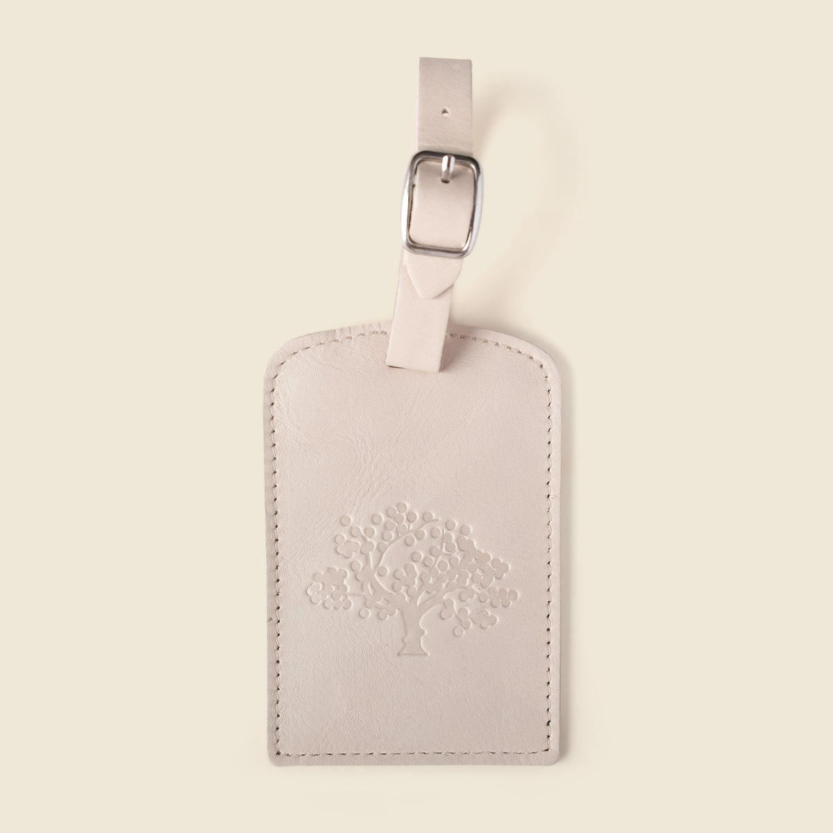 A pale blush pink leather luggage tag with a debossed tree design on the front, featuring a matching leather strap and silver metal buckle, set against a solid cream background.