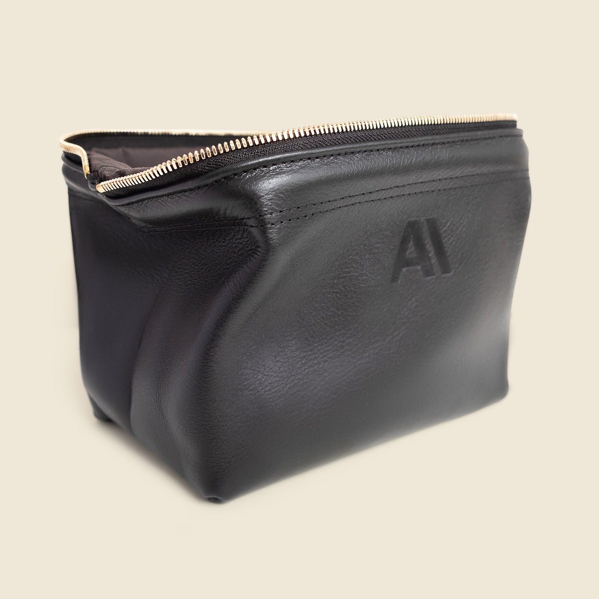 A black leather cosmetic pouch with a gold zipper and an embossed logo on the front, set against a solid light beige background.