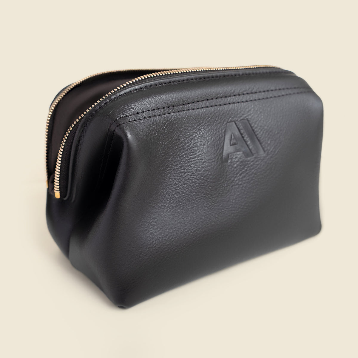 A black pebbled leather cosmetic pouch with a gold zipper and an embossed logo on the front, set against a solid light beige background.