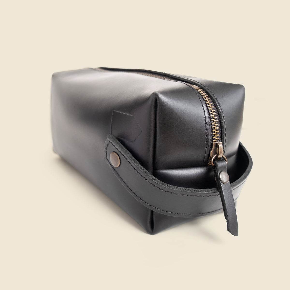 A black leather cosmetic pouch with a gold zipper and a matching leather wrist strap, shown on a solid light beige background.