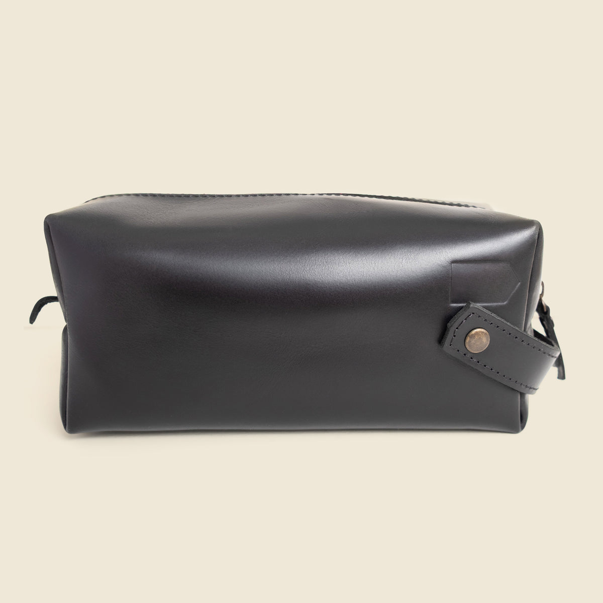 A black leather cosmetic pouch with a top zipper and a small leather strap detail with a gold-tone snap button, shown against a solid light beige background.