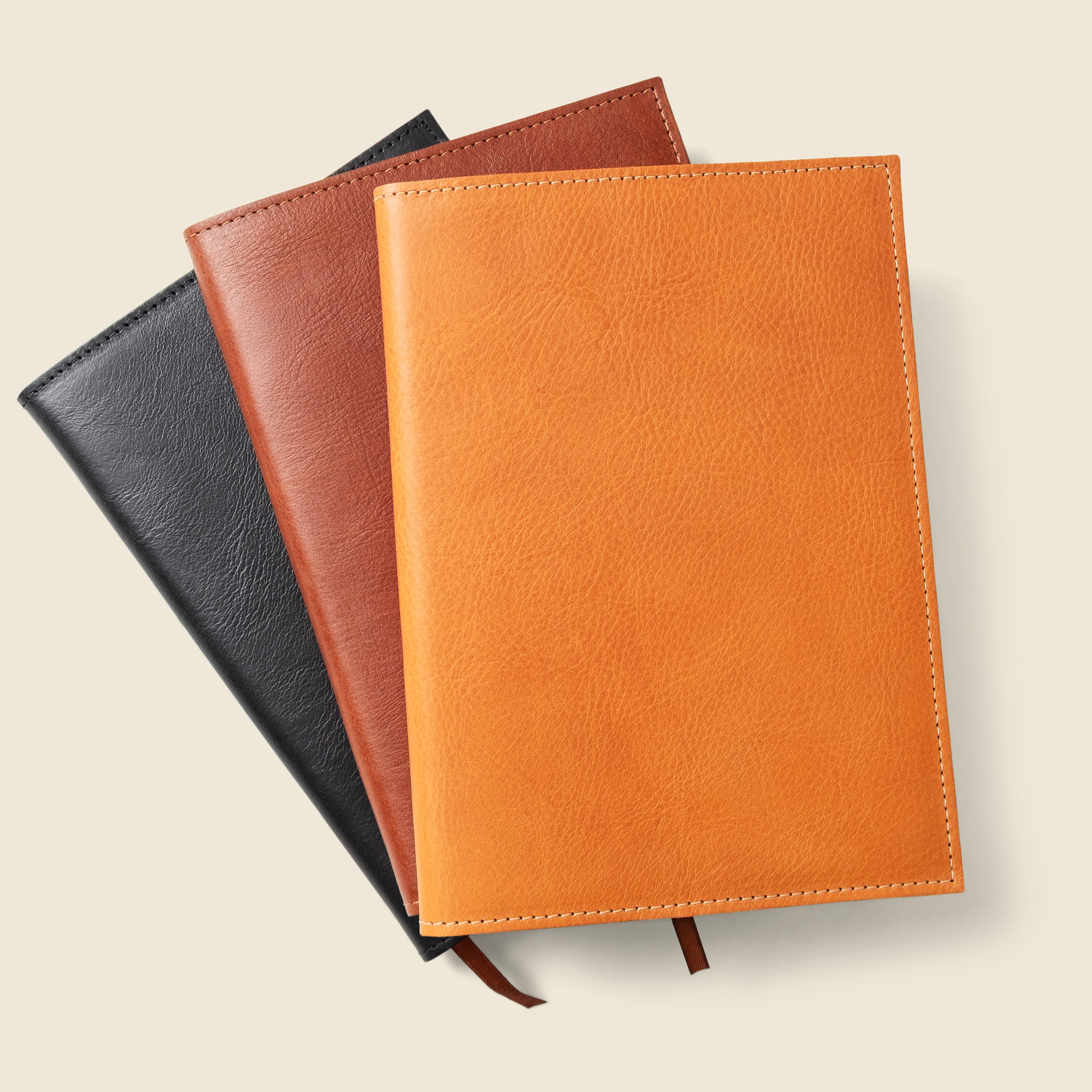 Three leather-bound notebooks in black, brown, and orange on a beige background