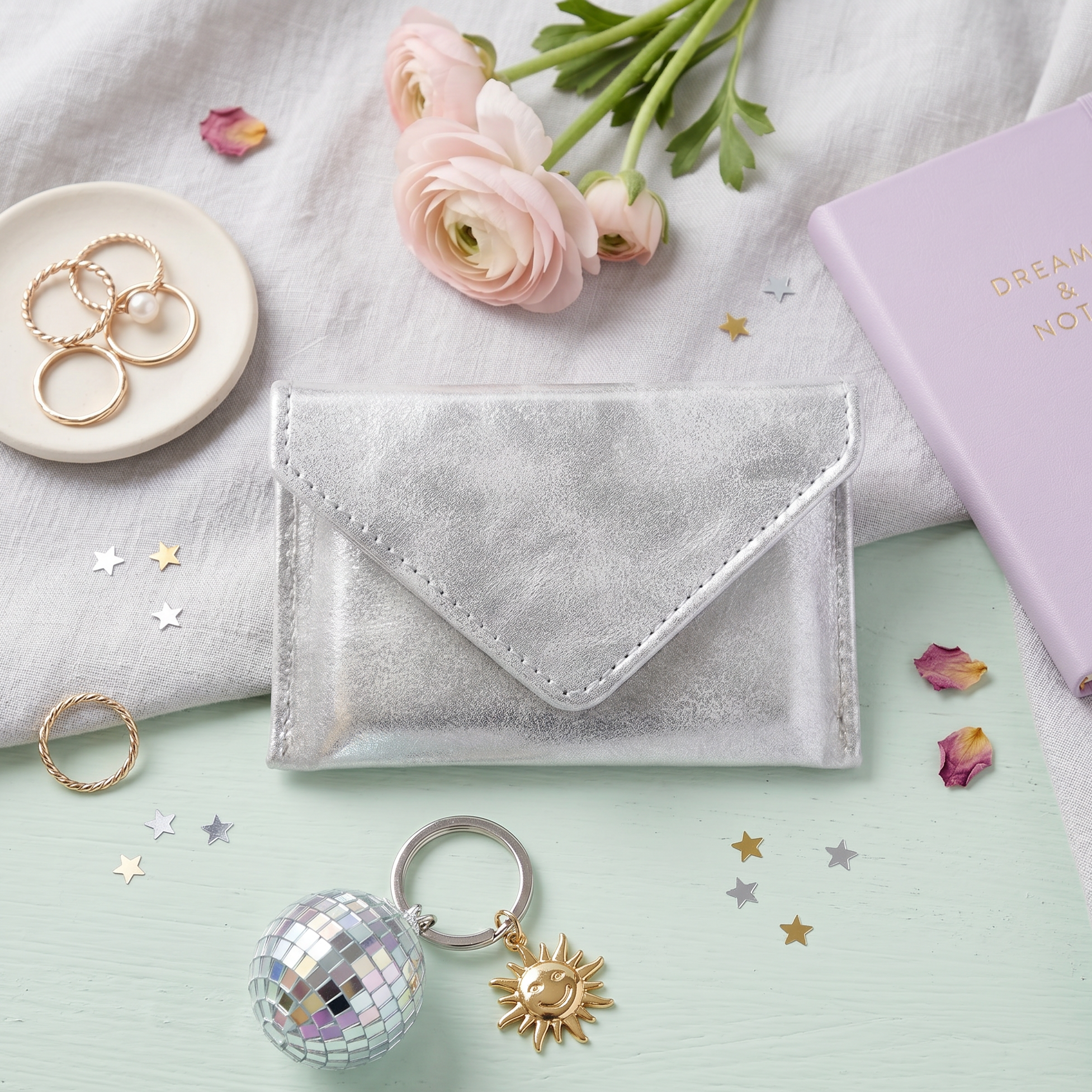 Silver glittery envelope, disco ball keychain, and gold rings on a light green surface with flowers and a notebook.